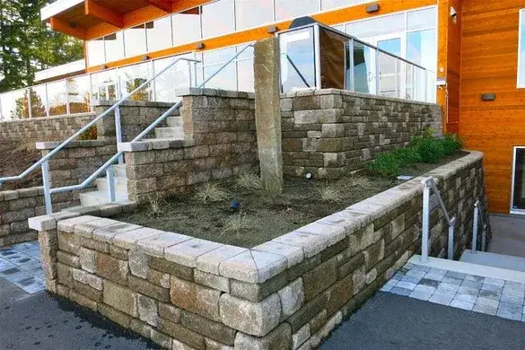 Tan segmental block retaining walls terracing a planted bed beside steps and metal handrails at the entrance of a modern cedar and glass building in Ballard