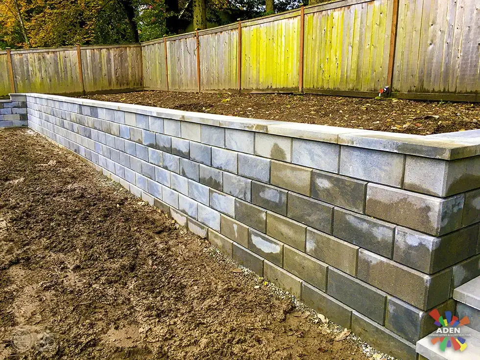 Long grey block retaining wall running the full length of a garden below a weathered timber boundary fence