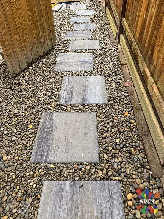 Three large grey stone slabs set as stepping stones in washed river rock between a timber fence and a raised bed