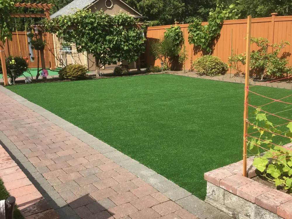 Rectangular artificial turf lawn bordered by red brick pavers, with a grape trellis and a raised brick planter alongside