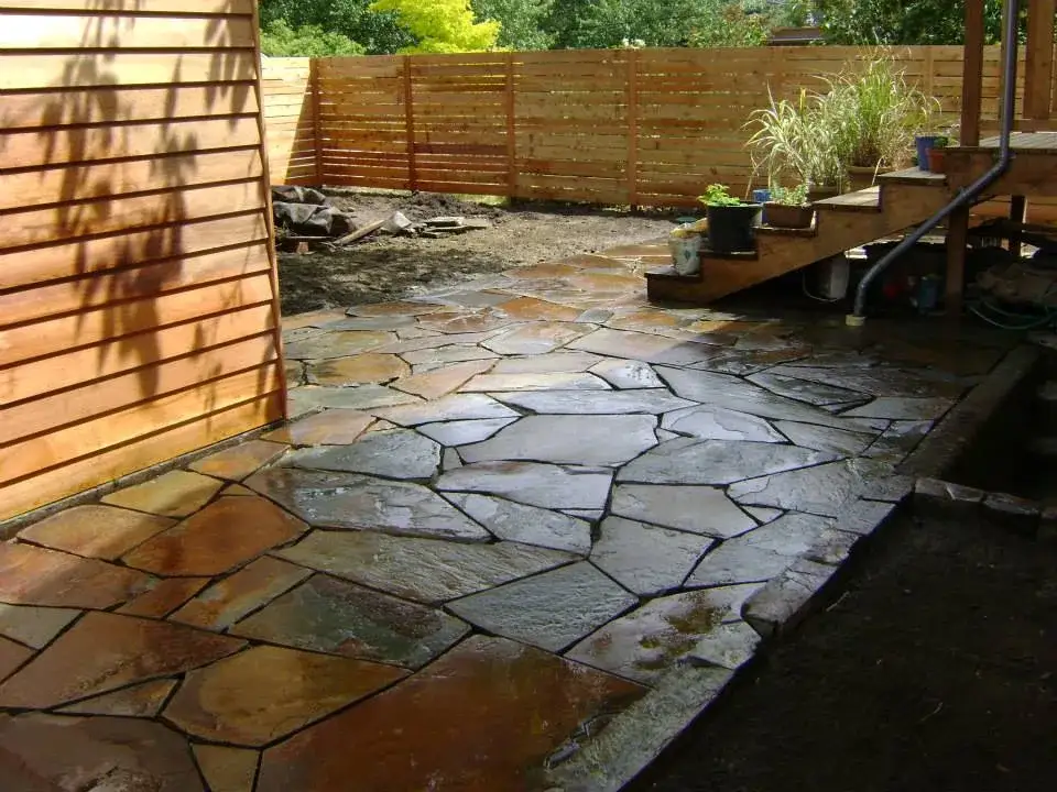 Wet irregular flagstone patio with dark joints in a small back garden enclosed by a horizontal cedar fence