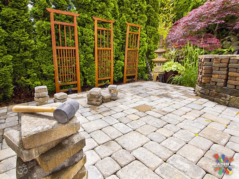 Partly finished pale paver patio with three cedar trellis screens behind it, stacked wall block and a rubber mallet in the foreground