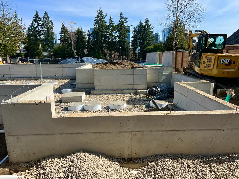 Excavator working alongside a house foundation on a levelled Bellevue site with drain rock and pipe runs laid out across the cut