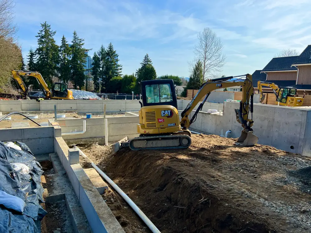 Excavator cutting and trenching across a levelled Bellevue site alongside a house foundation, with the city skyline in the distance