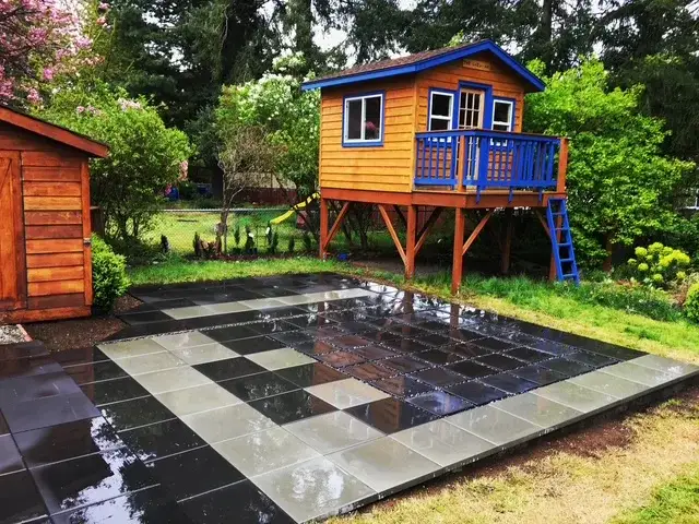 Chequerboard patio of large light and dark concrete slabs, wet with rain, beside a raised timber playhouse