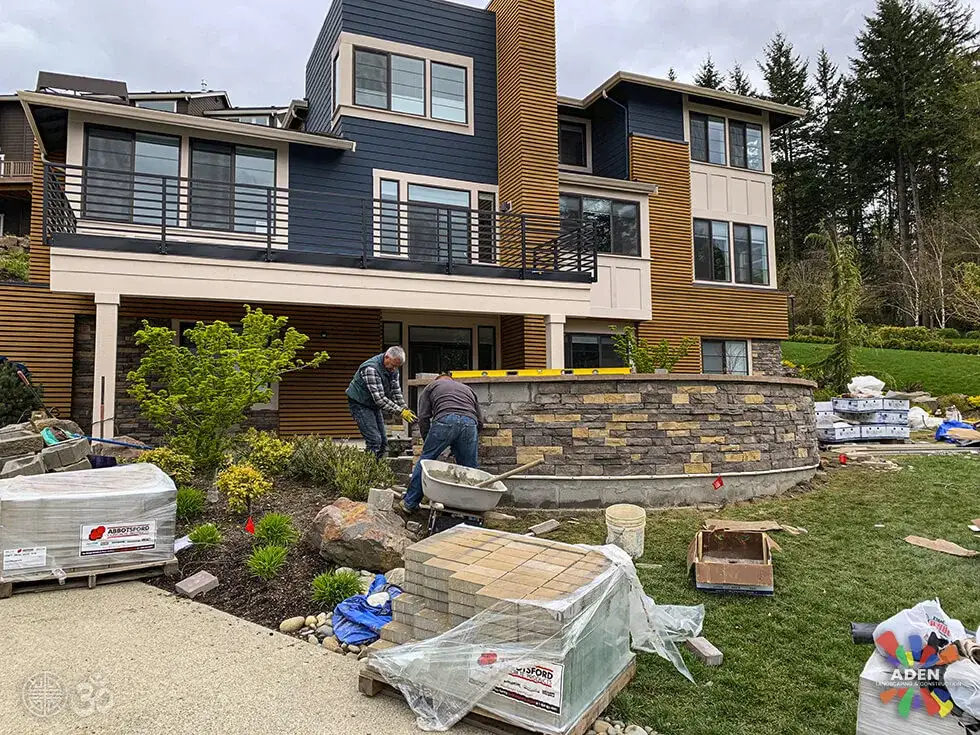 Two crew members laying stone veneer onto a curved front retaining wall outside a modern grey and timber house