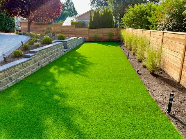 Artificial turf lawn beside a tan block retaining wall and new cedar fence, with ornamental grasses planted along the edge
