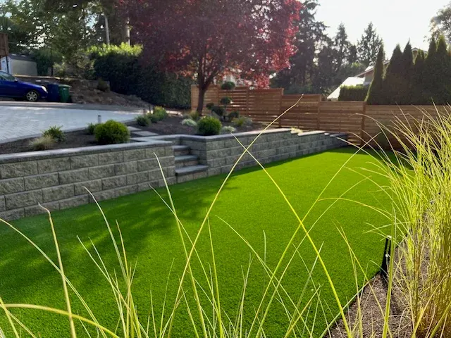 Block retaining wall with steps up to a paver terrace, seen across the turf lawn in late afternoon light