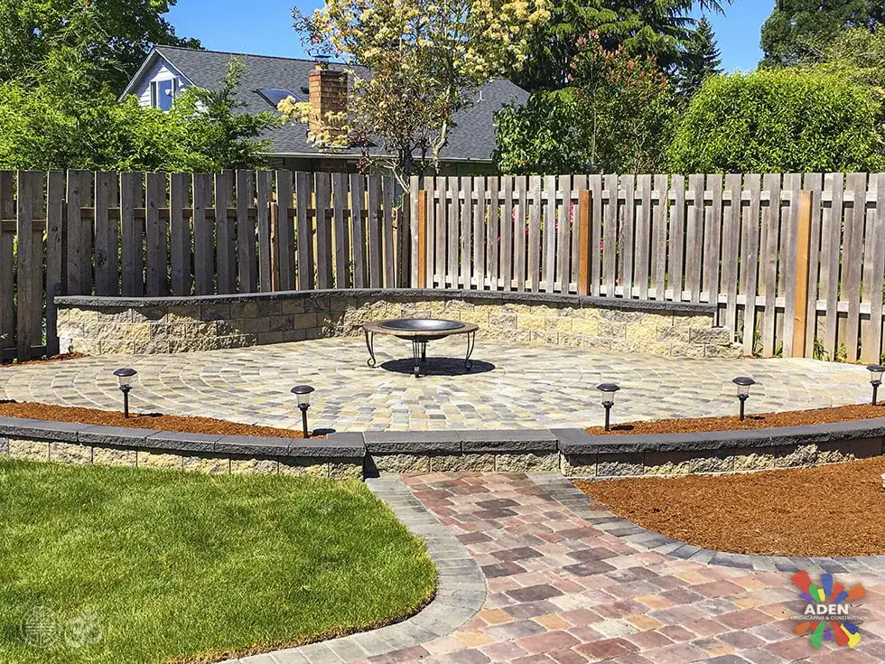 Paver terrace with a central stone fire pit and path lights, reached by a curving paver walkway across a lawn