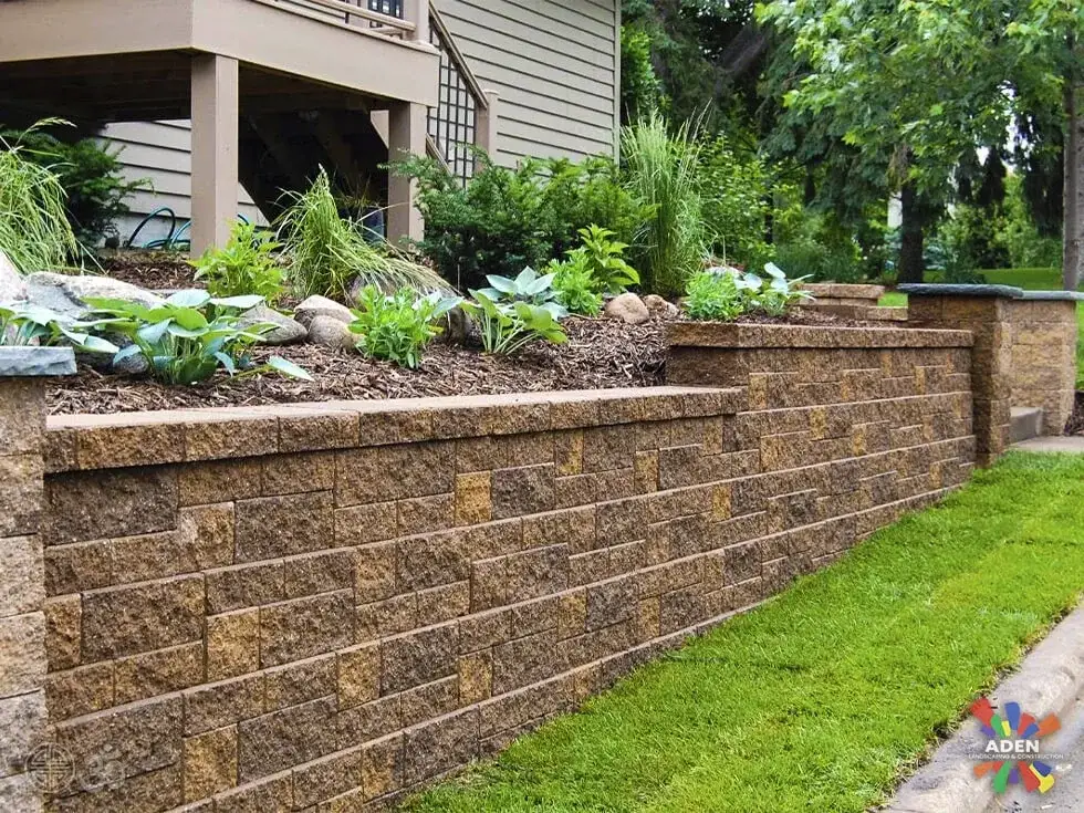 Tan split-face block retaining wall holding a planted bed of hostas and ornamental grasses above a level lawn