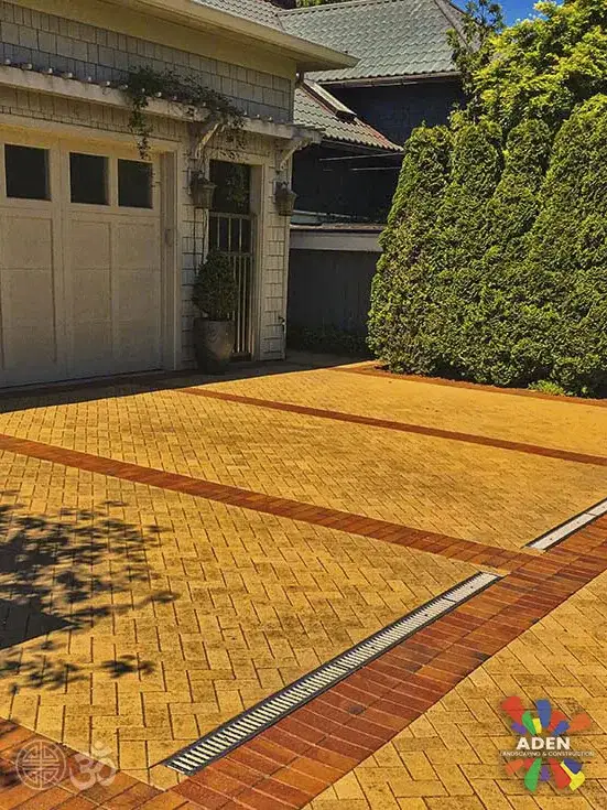 Tan paver driveway apron with a darker banded border running up to a garage door beside a tall clipped hedge