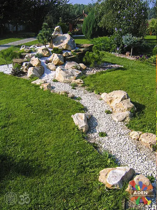 Dry creek bed of white gravel and set boulders curving through a lawn towards a rockery of larger stones