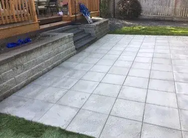 Large light-grey concrete slabs laid as a patio below an existing timber deck, against a low block retaining wall