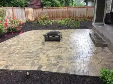 Tumbled paver patio with a black fire table, new planting beds and a cedar fence behind, wet after rain