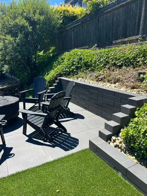 Charcoal block retaining wall with stepped returns holding a planted bank, edged with river rock above an artificial turf lawn and slab terrace