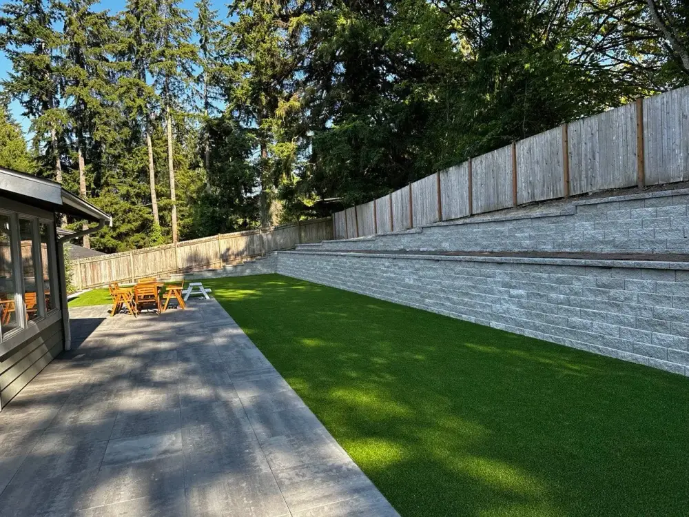 Large-format grey slab walkway beside a tiered block retaining wall with an artificial turf lawn on a sloped Magnolia lot