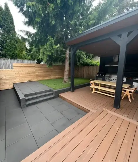 Covered outdoor kitchen and warm-toned deck beside a grey slab patio and turf lawn in a Magnolia garden