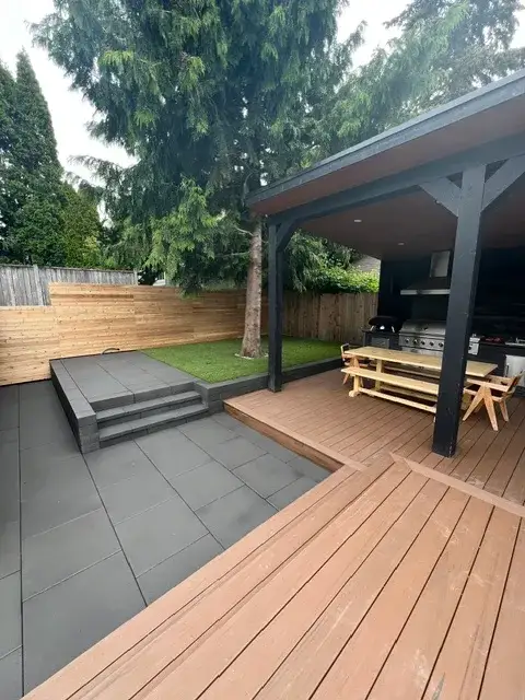 Covered outdoor kitchen and warm-toned deck beside a grey slab patio and turf lawn in a Magnolia garden