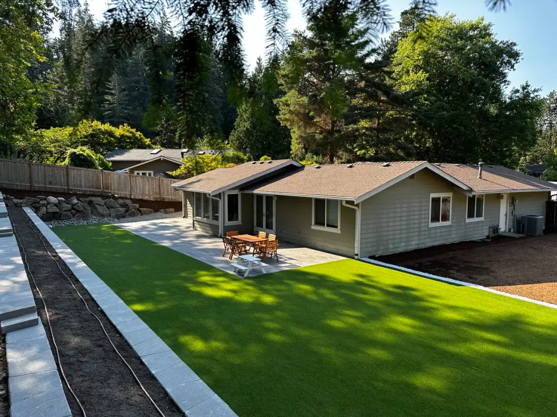 The same Magnolia garden finished: a paver terrace against the house with a dining set, a level artificial turf lawn, and a block wall edge with a boulder rockery beyond