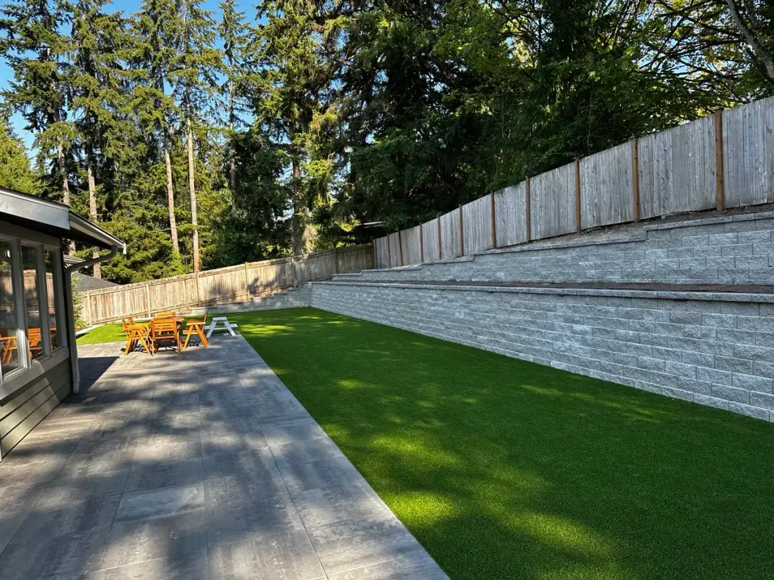 The same view finished: a tall grey block retaining wall stepping up in terraced courses along the boundary, above a level artificial turf lawn and a large-format paver terrace
