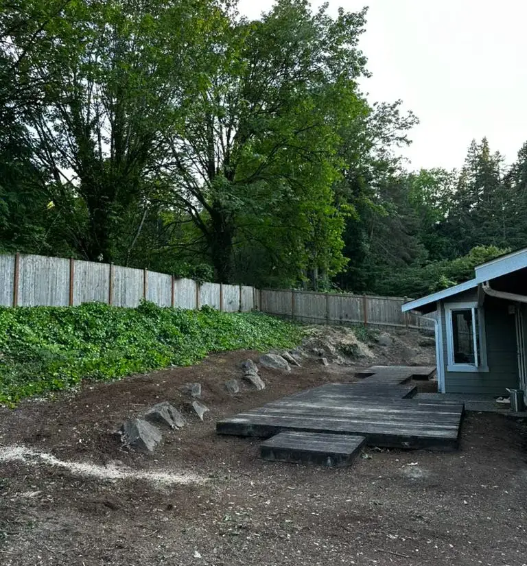 Magnolia back garden before work: bare soil, a weathered timber deck platform against the house, and an ivy-covered bank held back by loose boulders under a cedar fence