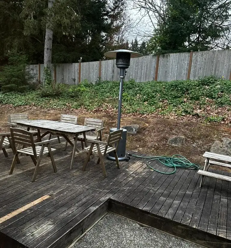 The same garden the previous winter: a dark weathered timber deck with a patio heater and folding furniture, backed by an ivy bank and a leaning cedar fence