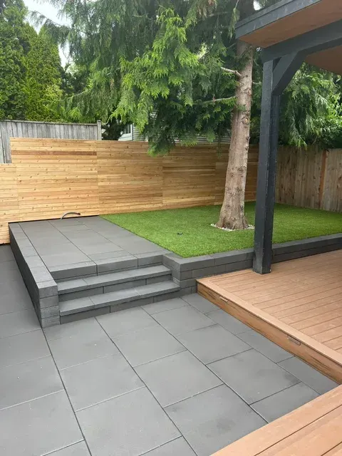 Grey slab patio with a stepped edge rising to an artificial turf lawn beneath a mature fir, enclosed by horizontal cedar screen fencing