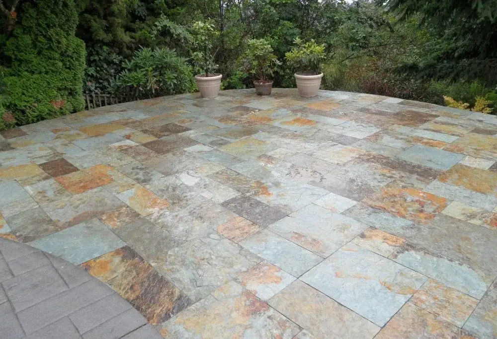 Large slate terrace laid in a random pattern of rust, green and grey tiles, with potted shrubs along a clipped hedge