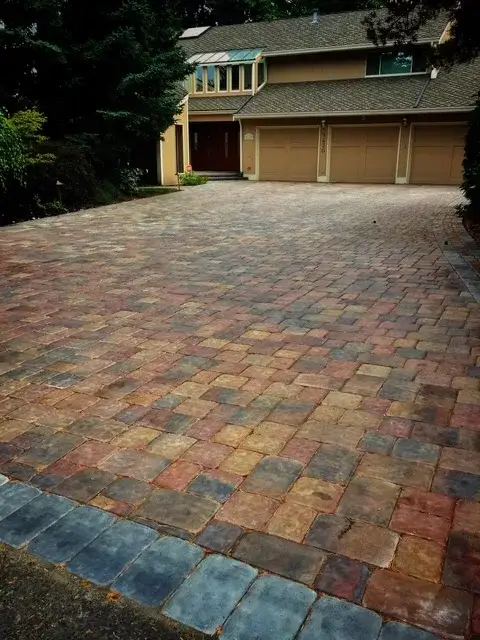 Wide tumbled paver driveway in mixed autumn tones running up to a three-bay garage on a wooded lot