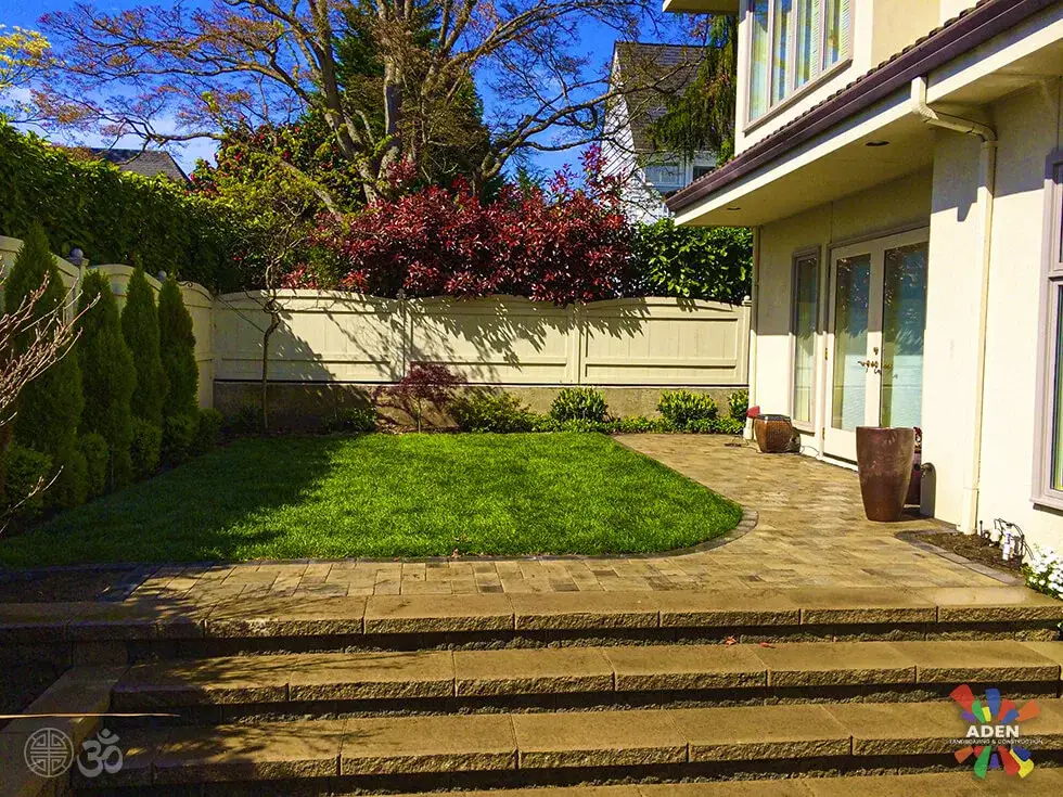 Wide paver steps leading down from a house to a paved terrace with a curved lawn edge and clipped hedging behind
