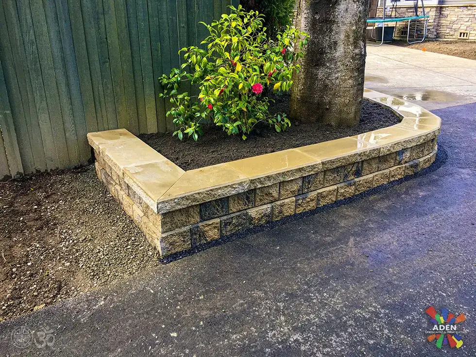 Low cut-stone wall with a wide cap holding a mulched planting bed beside a wet asphalt driveway
