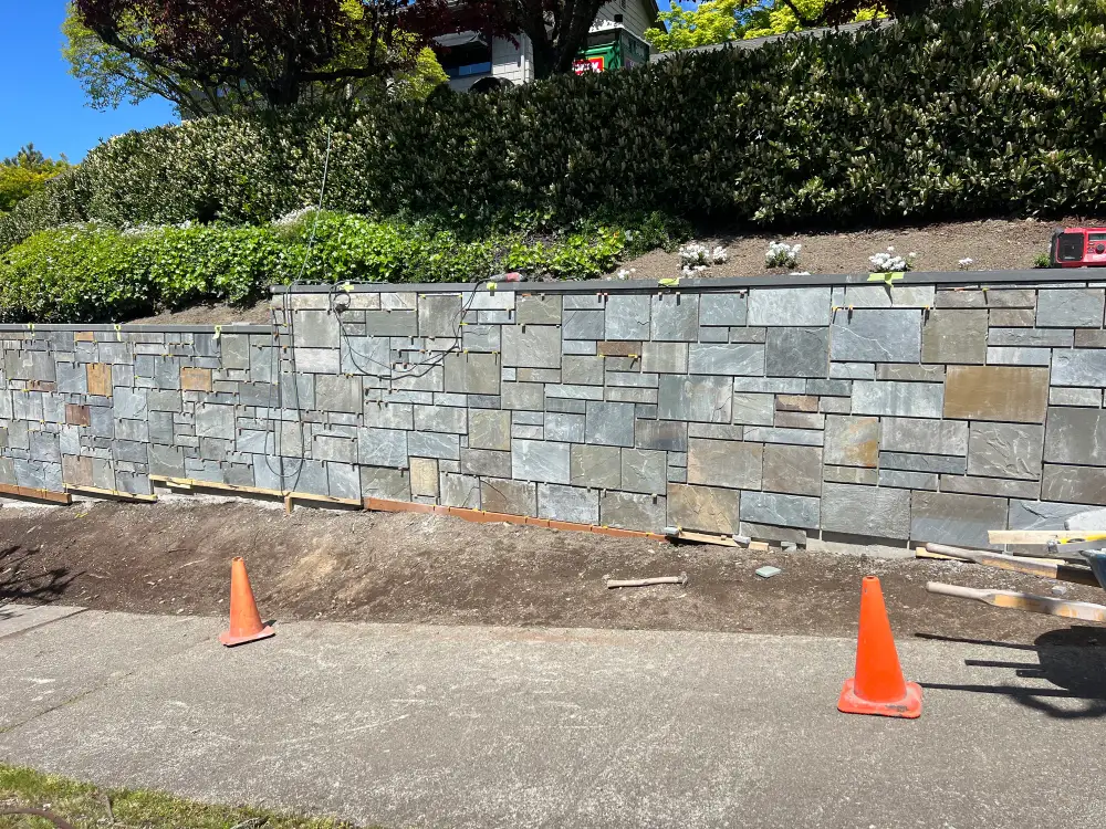Long cut-stone veneer retaining wall under construction along a street frontage with traffic cones set out in front of it
