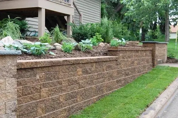 Long tan split-face block retaining wall with a flat capping course holding a raised bed of hostas, grasses and boulders beside a lawn