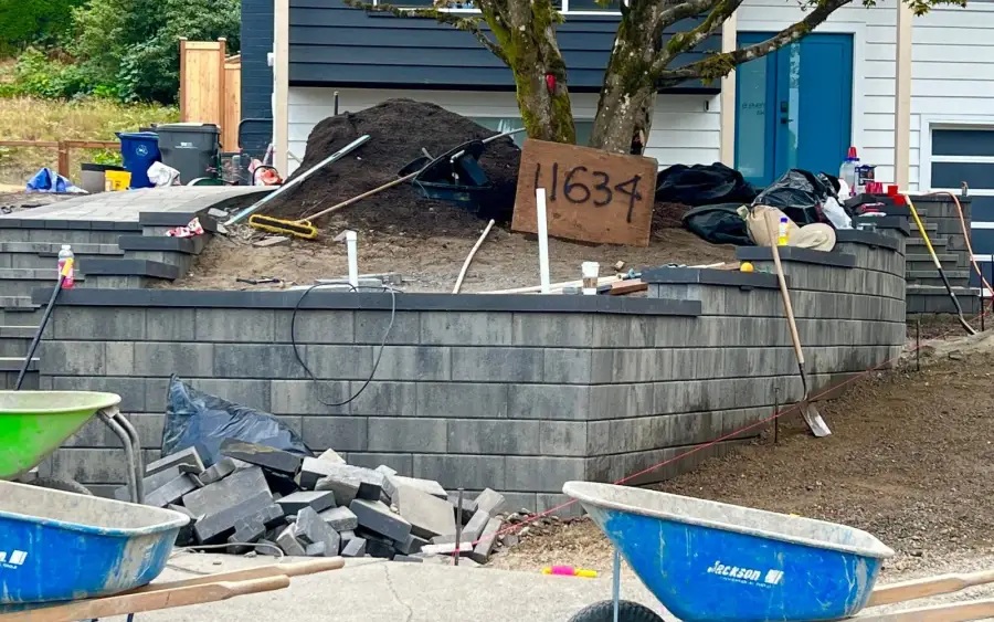 Charcoal block retaining wall under construction with capping units, wheelbarrows and stacked material on site