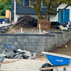 Charcoal block retaining wall under construction with capping units, wheelbarrows and stacked material on site