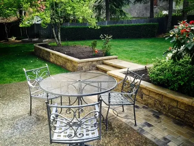 Low stone seat wall and raised planting bed with wide stone steps up to a lawn, and a table and chairs on the paved terrace