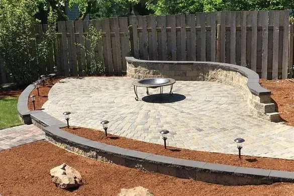 Circular paver patio with a contrasting inner ring under a fire bowl, enclosed by a capped curved seat wall with path lights along the mulched bed edge