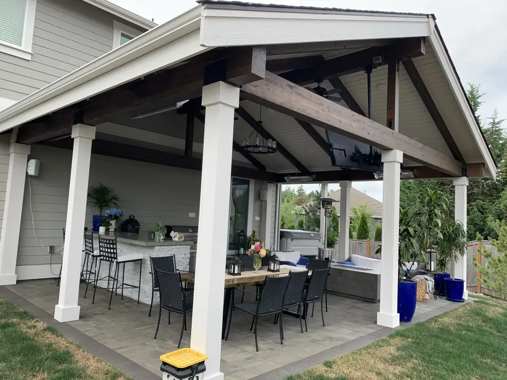 Covered outdoor pavilion with a gabled roof, dark stained beams and white posts over a paver terrace, with a stone-faced outdoor kitchen bar, dining table and hot tub beneath it