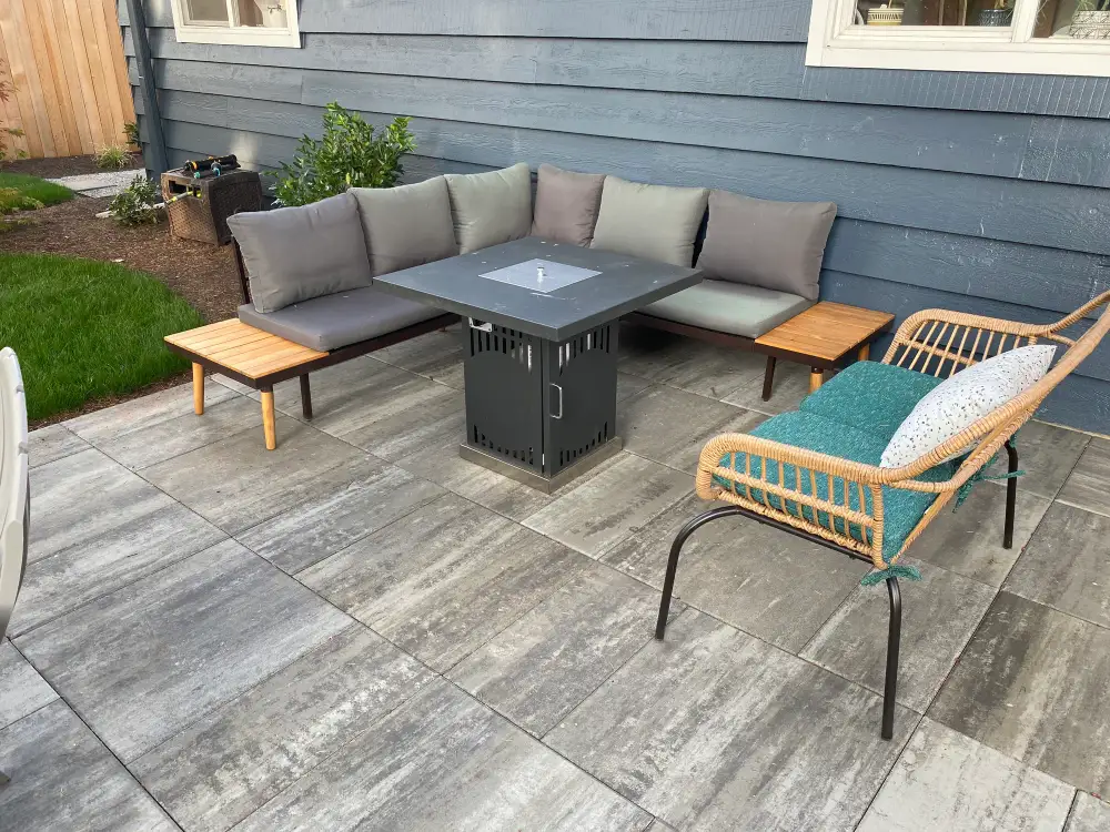 Large-format grey slab patio against a blue-grey clapboard house, with a corner sofa, a square gas fire table and a rattan chair on it