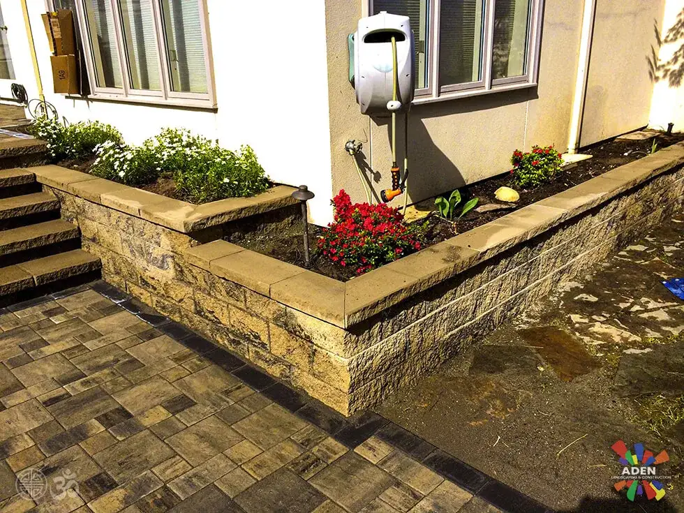 Stone-faced raised planting beds wrapping the corner of a cream stucco house, freshly planted with red flowers