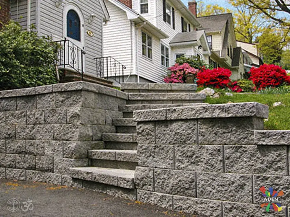 Grey split-face block retaining wall with integrated steps in front of a white clapboard house, with flowering shrubs above it
