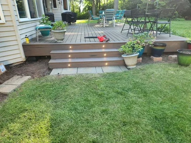 The same deck seen from the lawn, its lit riser steps landing on a paver strip set into fresh turf, with a dining set on the deck above