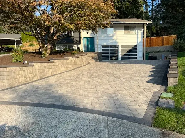 Wide grey paver driveway running up to a garage, with terraced block retaining walls and planting beds on both sides
