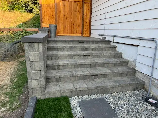 Wide grey block steps with a capped pier running up the side of a house to a new cedar gate, with a rock drainage strip alongside