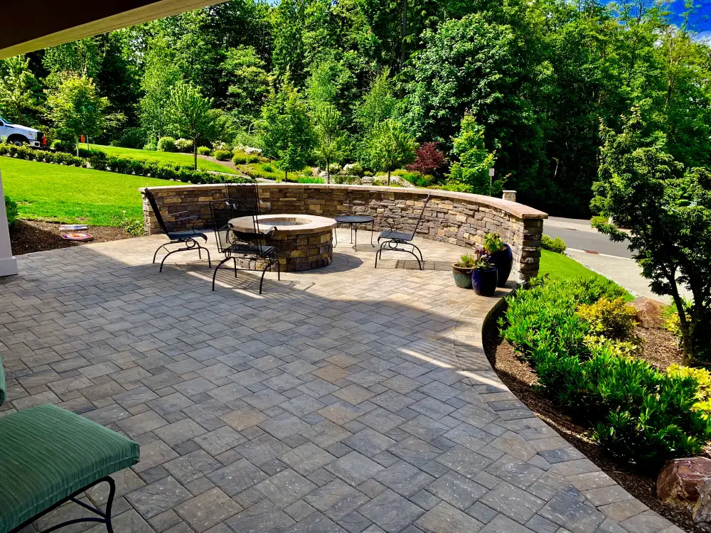 Long curved stone seat wall around a paver terrace with a round fire table and metal chairs, backed by clipped hedging