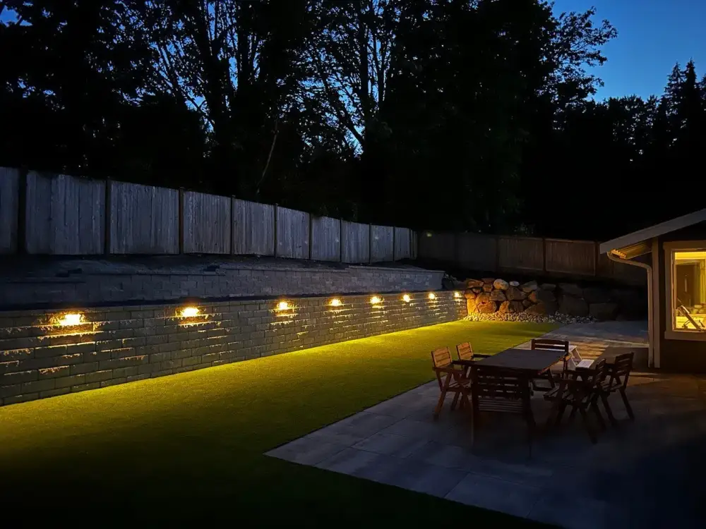Retaining wall washed with warm landscape lighting at night above a lit patio and dining set