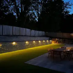 Retaining wall washed with warm landscape lighting at night above a lit patio and dining set