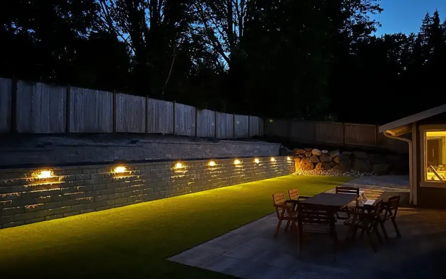 Retaining wall washed with warm landscape lighting at night above a lit patio and dining set