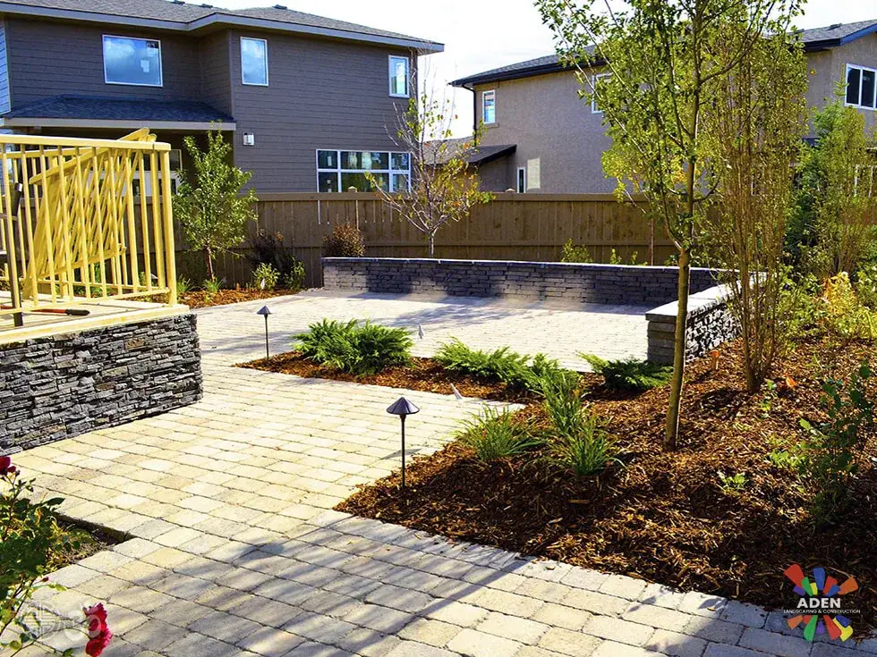 Pale paver patio with a stone-faced seat wall, freshly planted beds and a yellow railing beside a modern house