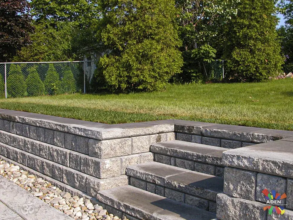 Grey block retaining wall with built-in steps and a course of white drainage rock along its base beside a lawn